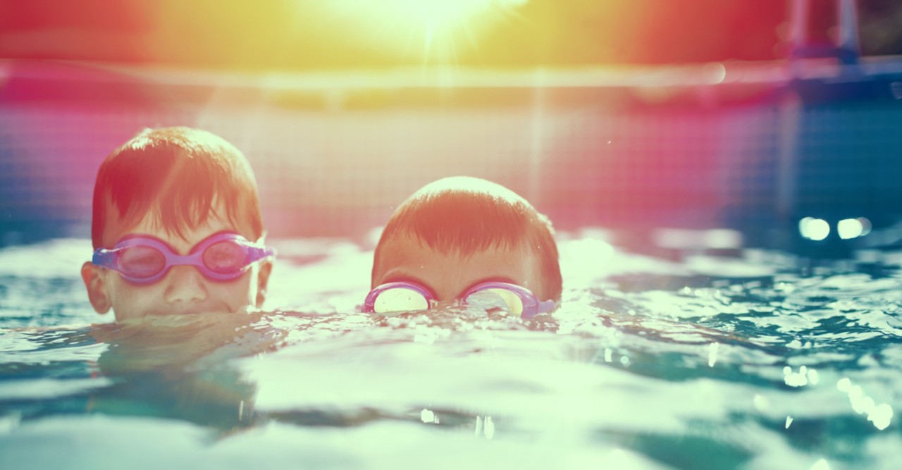 两个戴着护目镜的小孩在夕阳下的泳池里游泳”title=