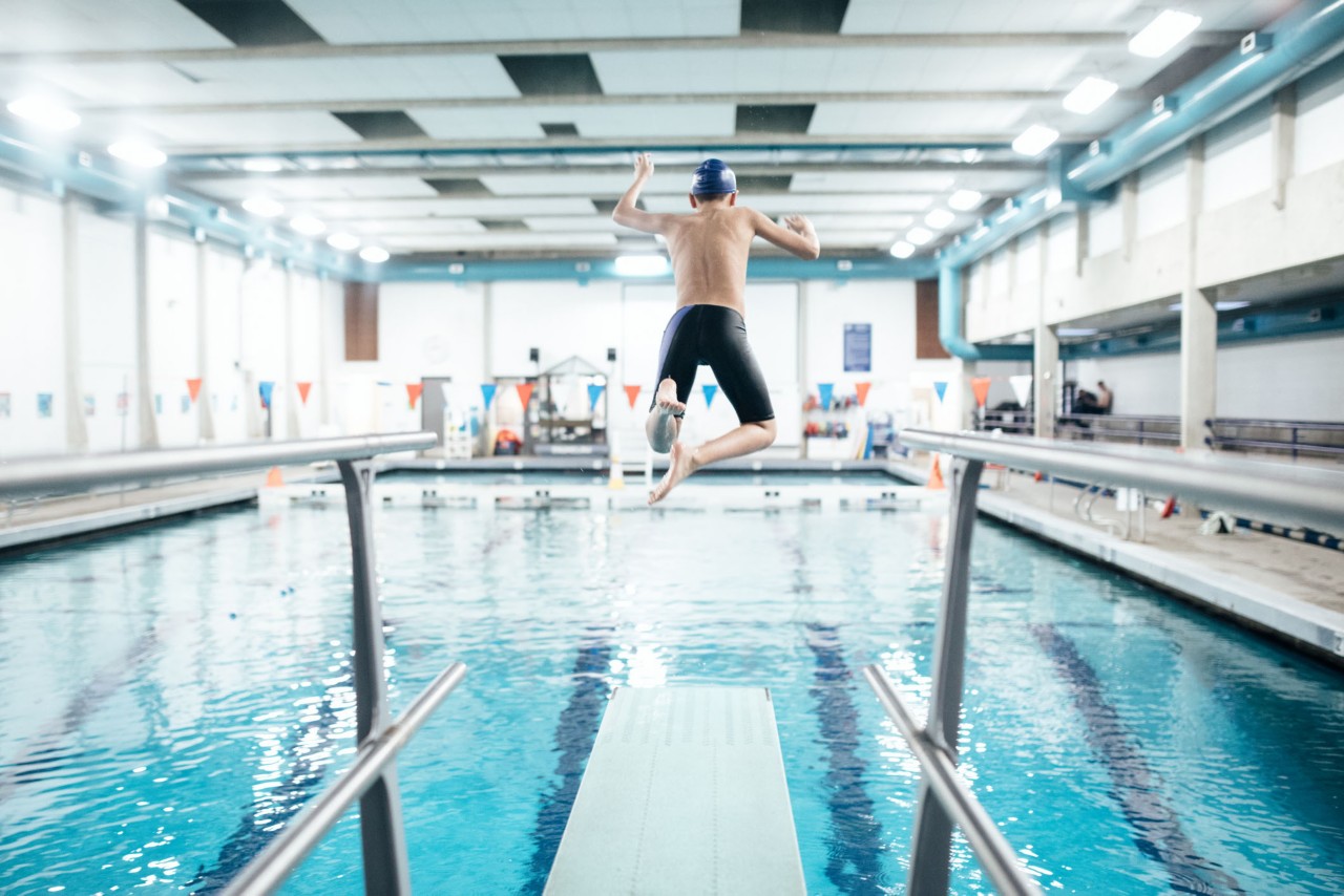 一个12岁的男孩在游泳池里享受着从跳板上跳入水中的时光。健康积极的生活方式也很有趣。＂title=