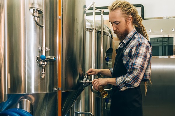 啤酒厂的酿酒师将啤酒从啤酒厂的水箱中倒入玻璃杯中＂class=