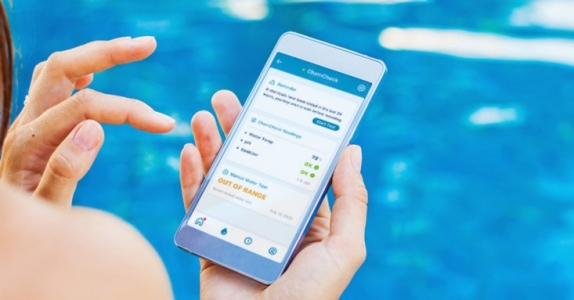 一名女子坐在泳池边，用手机展示水的化学反应，泳池自动化＂height=