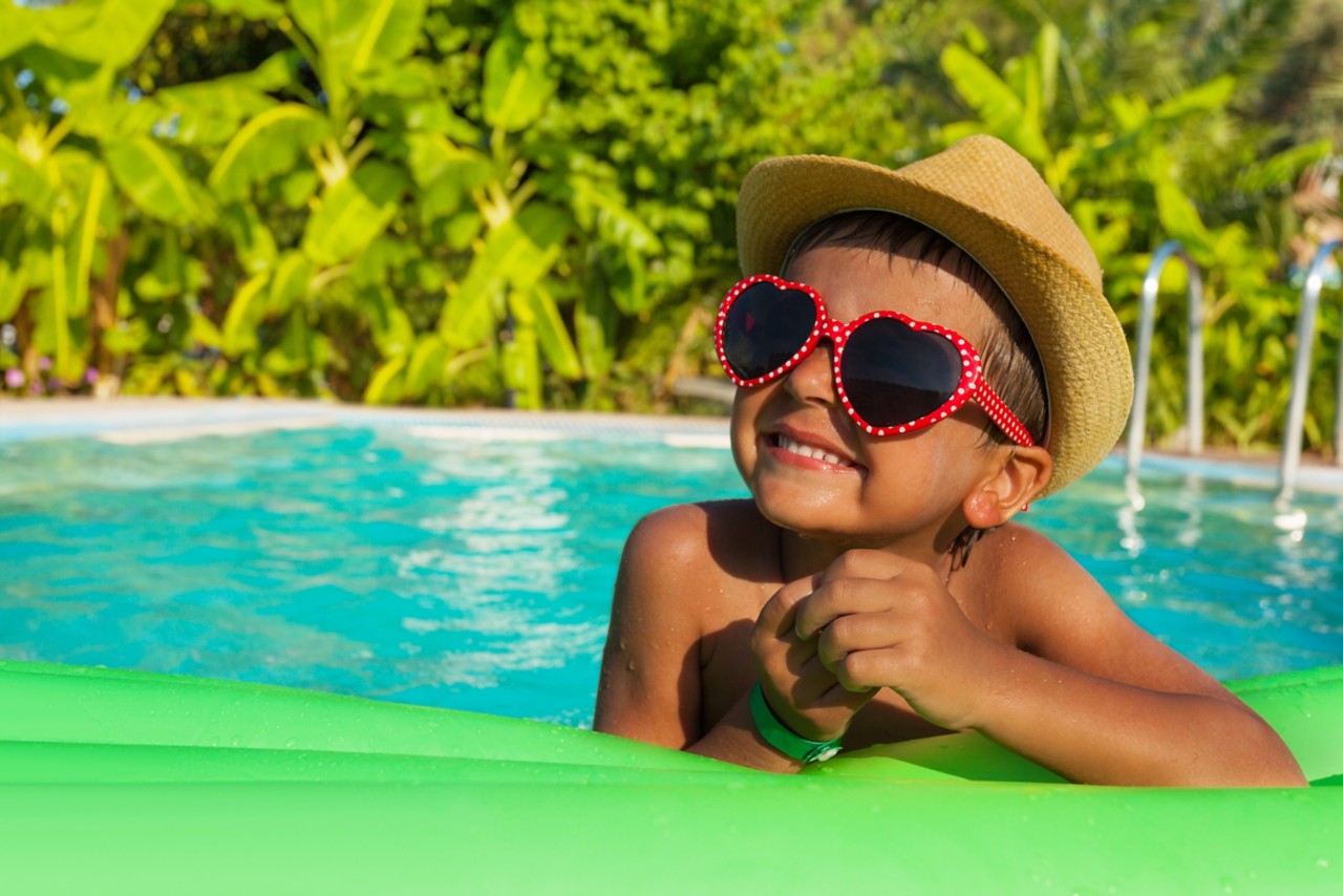 可爱的男孩在心形太阳镜和帽子上的绿色充气床充气在游泳池外的夏天