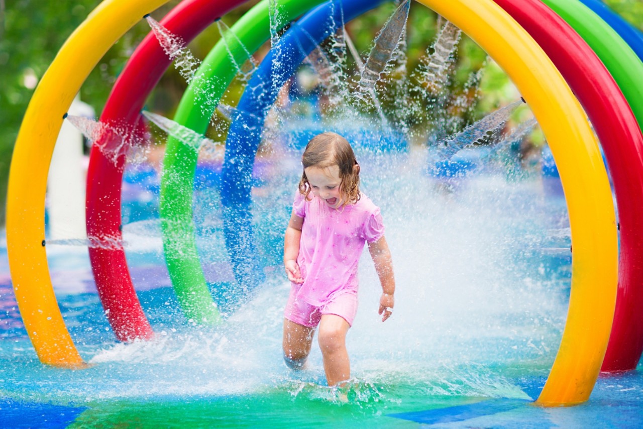 快乐的小蹒跚学步的女孩通过一个充气玩具洒水车有乐趣的水飞溅在游泳池享受一日游到水上游乐园在夏季家庭度假＂title=