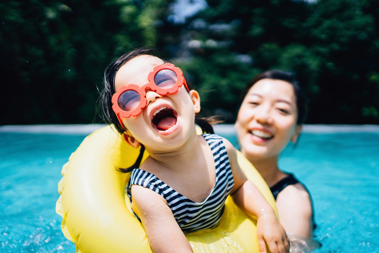 戴着墨镜的快乐蹒跚学步的小女孩快乐地微笑着，享受着与母亲在游泳池里嬉戏的家庭纽带时光