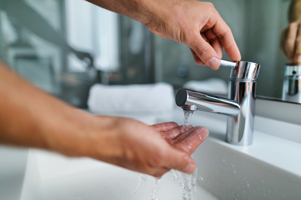 一名男子在家中浴室洗手池洗手，用手触摸自来水检查温度。从水槽的水龙头流出热水的手指特写;Adobe股票，188032490＂class=